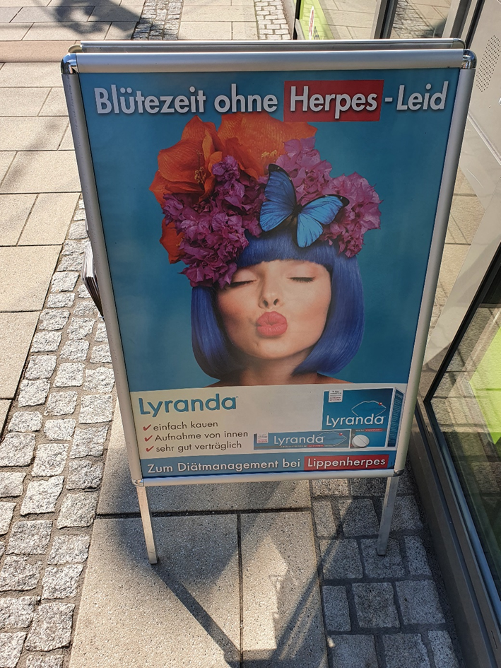Werbeaufsteller mit dem Gesicht einer Frau mit blauen Haaren und dem Titel "Blütezeit ohne Herpes-Leid"