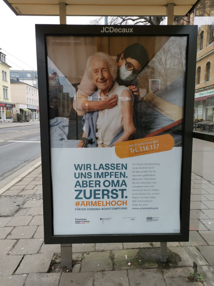 Werbeplakat an einer Haltestelle von einer älteren Frau, die geimpft wurde mit dem Titel "Wir lassen uns impfen. Aber Oma zuerst. Ärmelhoch