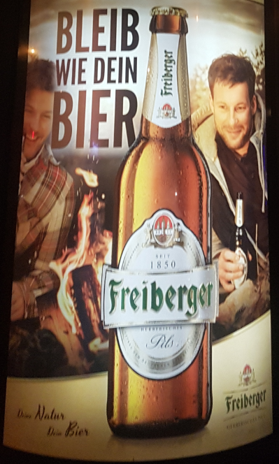 Selbes Werbeplakat der Biermarke "Freiberger" mit dem Titel "Bleib wie dein Bier" mit zwei Männern an einem Lagerfeuer