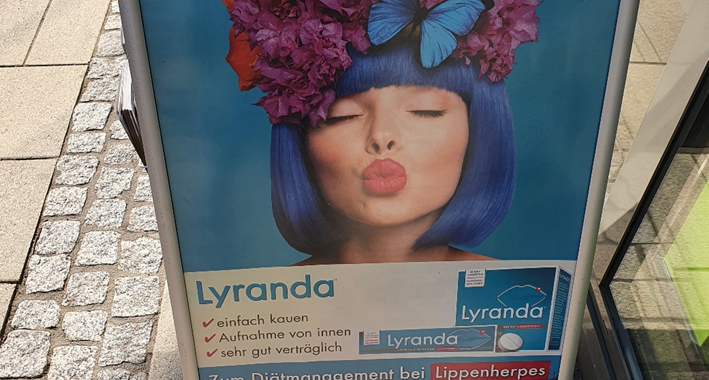 Werbeaufsteller mit dem Gesicht einer Frau mit blauen Haaren und dem Titel "Blütezeit ohne Herpes-Leid"