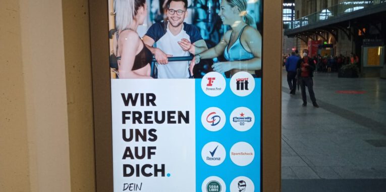 Foto eines Werbeplakates mit dem Titel "Wir freuen uns auf dich. Dein Fitness-Studio". Im Vordergrund unterhalten sich in einem Fitness-Studio zwei leicht bekleidete Frauen mit einem Mann.