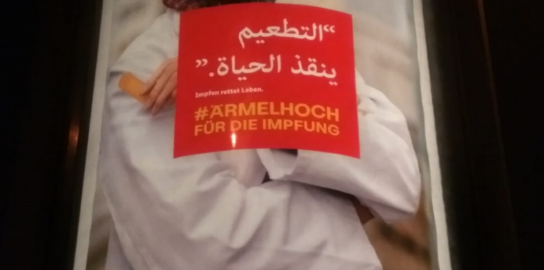 Bild eines Werbeplaktes mit einer jungen Frau mit Kopftuch, die sich ein Pflaster an den Oberarm hält mit dem Titel: "Impfen rettet leben. #Ärmelhoch für die Impfung"