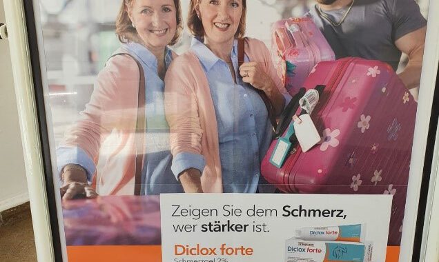 Foto eines Werbeplakat von Ratiopharm mit zwei sich stark ähnelnden, lächelnden Frauen und einem bärtigen Mann rechts daneben, der zwei rosa Koffer in den Händen hält.