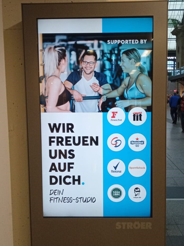 Foto eines Werbeplakates mit dem Titel "Wir freuen uns auf dich. Dein Fitness-Studio". Im Vordergrund unterhalten sich in einem Fitness-Studio zwei leicht bekleidete Frauen mit einem Mann.