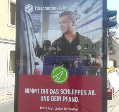 Werbeplakat von Flaschenpost.de auf dem ein junger Mann eine Getränkekiste in ein Fahrzeug läd.